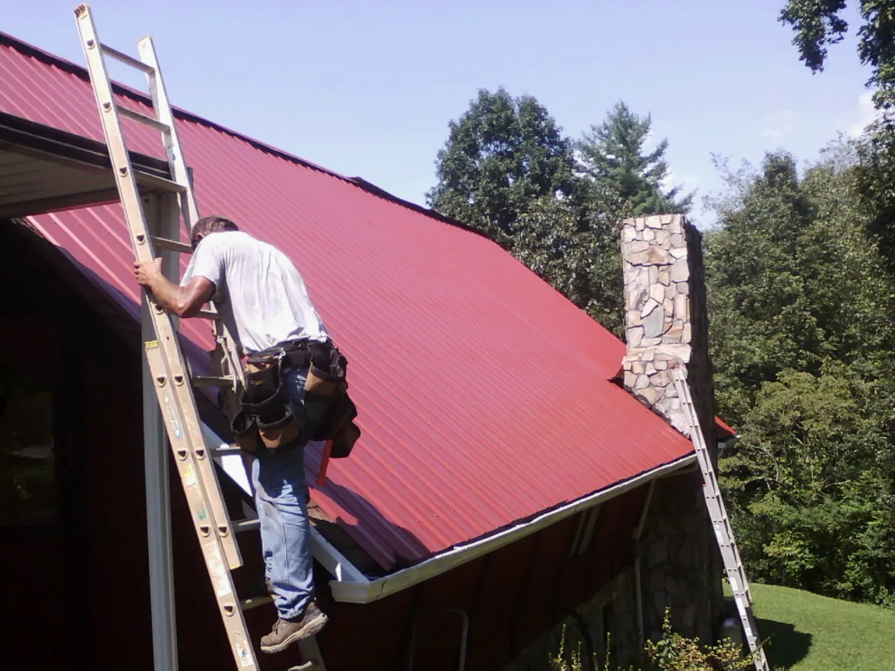 Roofer on a ladder beside a red metal roof and stone chimney — Soffit & Fascia Repair by Roofing Brothers in Keyesport